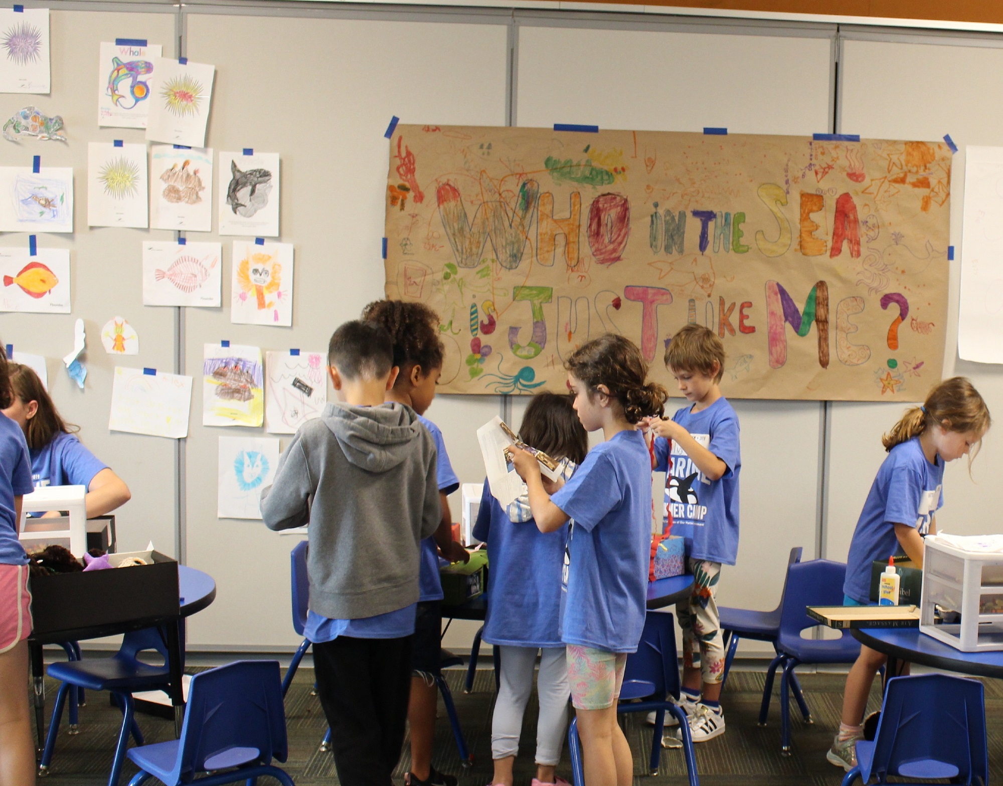 Several children making arts and crafts under a large, handmade paper banner that reads "Who in the sea is just like me?"