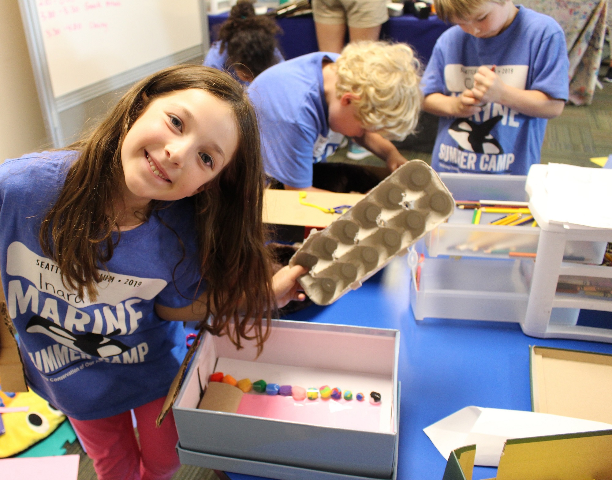 A child in a Marine Summer Camp shirt holding an empty egg carton and smiling.