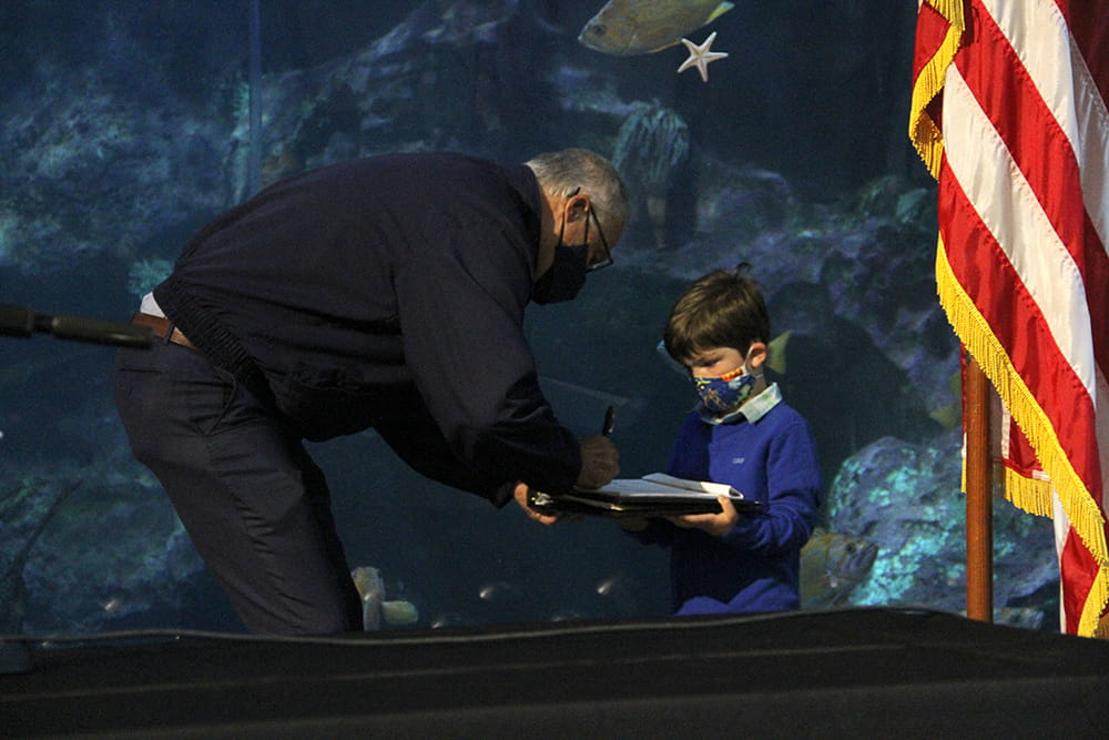 Governor Jay Inslee signing a document on a clipboard held by a young boy.