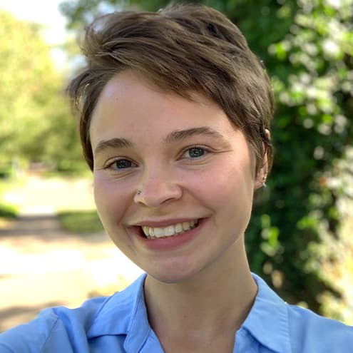 Jules Rader smiling for a photo. They have short brown hair and they are wearing a light blue shirt.