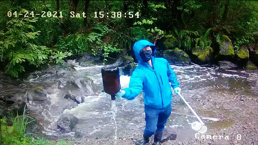 Dr. Zhenyu Tian conducts field research next to a river within a forest location, holding a metal pole with a plastic bottle attached in one hand, and emptying a large bottle of water in the other hand.