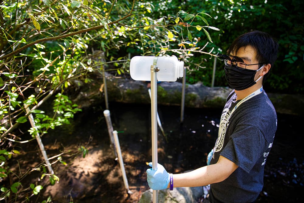 Dr. Zhenyu Tian conducts field research while standing in a forested location next to a river, holding a metal pole attached to a plastic bottle.