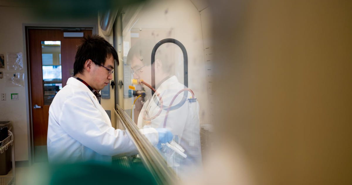 Dr. Zhenyu Tian, wearing a lab coat, stands in a laboratory while conducting work at a lab station behind a large glass panel.