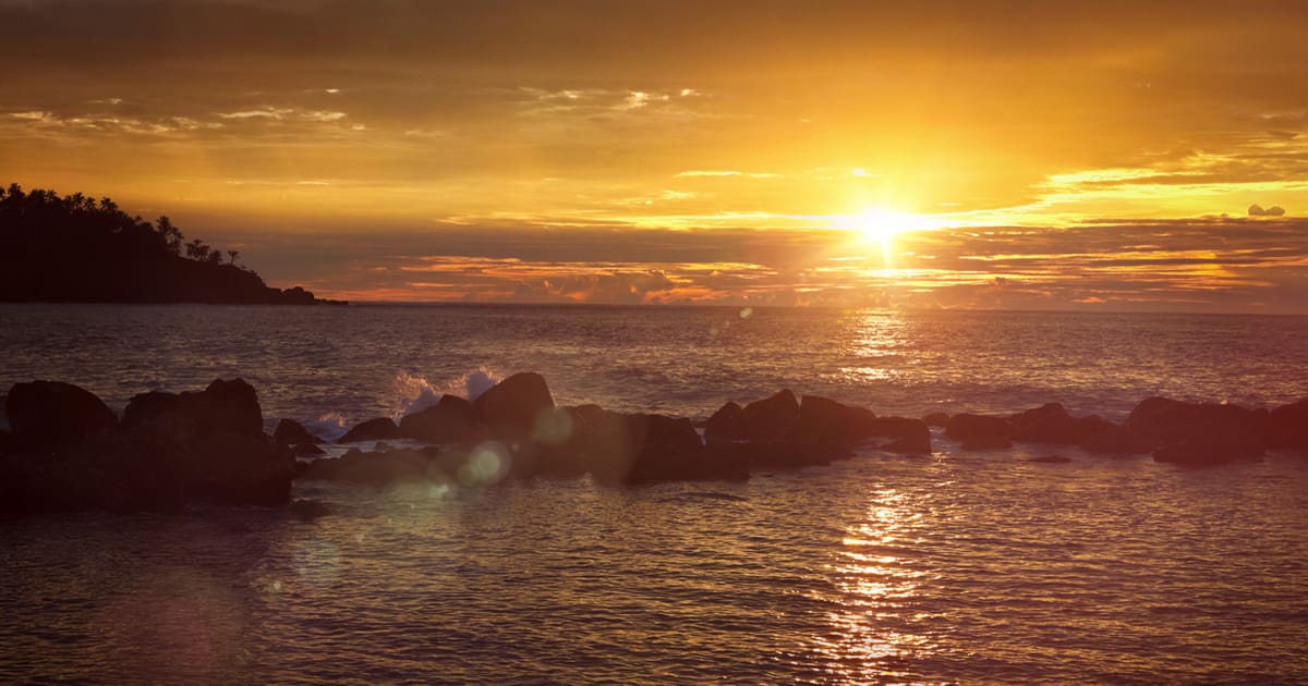 Waves crashing against a series of small rocks along an ocean shoreline during sunset.