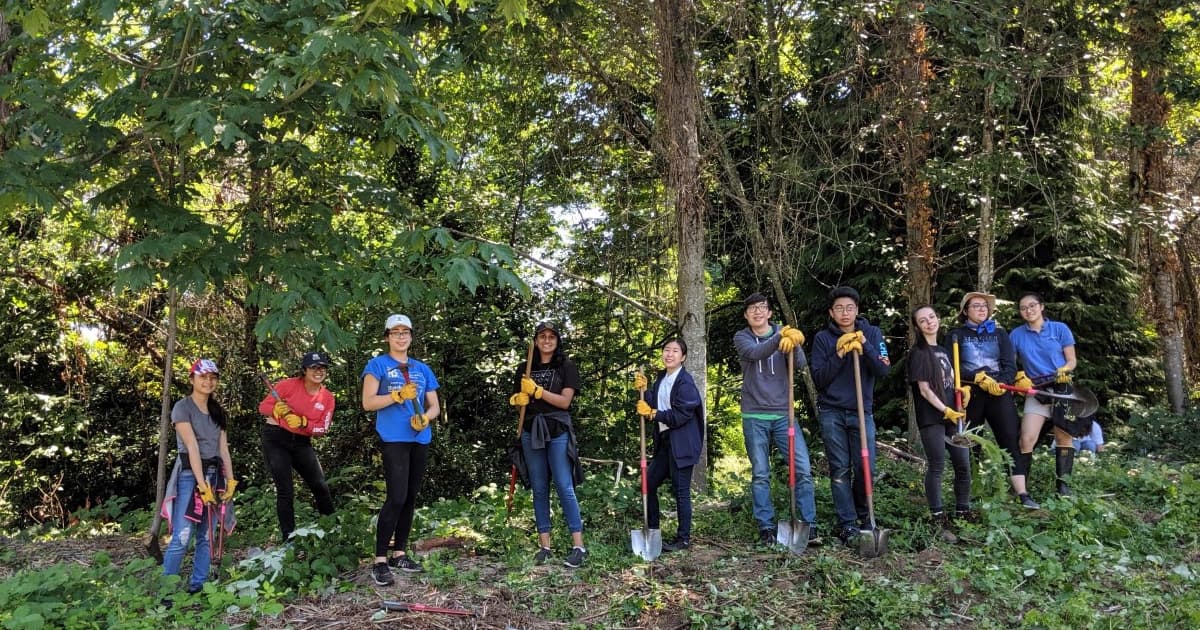 A group of 10 teenagers standing in a forested area holding shovels and other tools.
