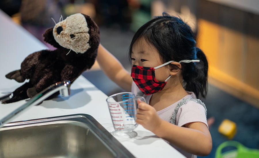 A young girl holding a stuffed sea otter toy and a measuring cup, approaching a play sink in Seattle Aquarium's Caring Cove play space.