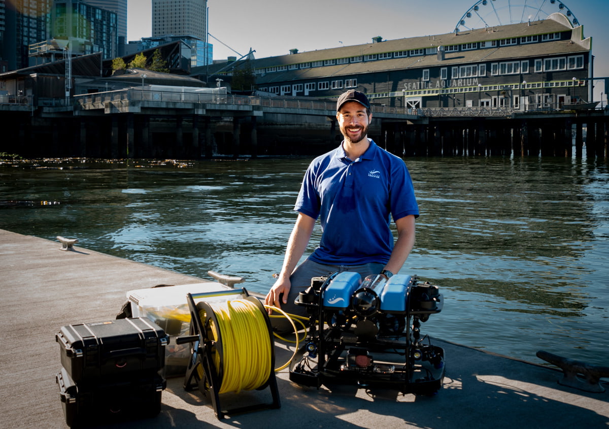 Dr. Zachary Randell poses while kneeling on a dock next to ROV Nereo and other equipment used for underwater research.