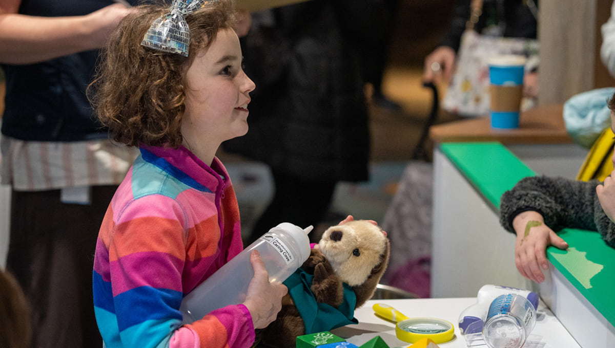 A young girl in a colorful, striped shirt "feeding" a stuffed otter with a plastic bottle.