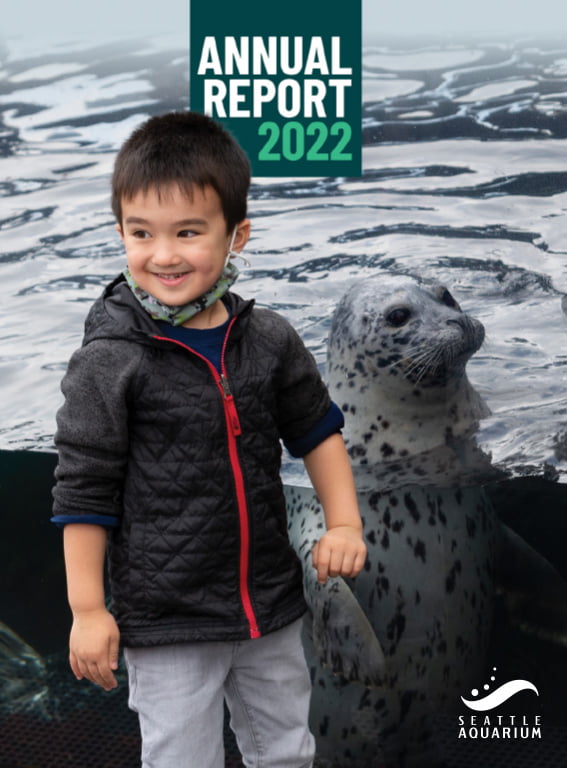 A photo of a young boy standing in front of the harbor seal habitat; a harbor seal floats vertically behind him. A small banner at the top reads "Annual Report 2022." The Seattle Aquarium logo is in the bottom right corner.