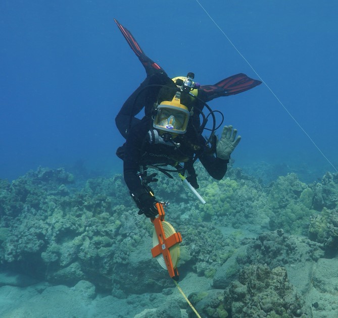 Amy Olsen waving at the camera while diving underwater. She is wearing full SCUBA gear and holding a round tape measurer.