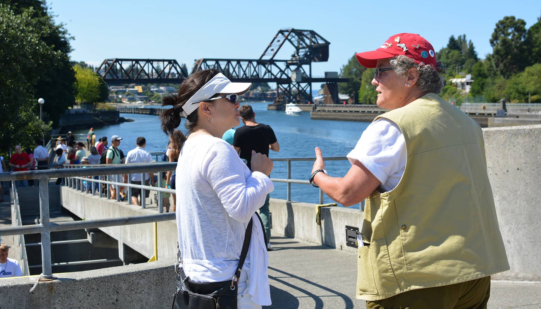 A volunteer wearing a beige vest and a red hat speaks to a person on the edge of the Cedar River.