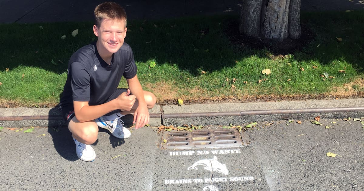 Lucas. C kneeling and giving a thumbs up next to a storm drain alongside a street in Seattle. The storm drain has been stenciled with the words "dump no waste, drains to Puget Sound".