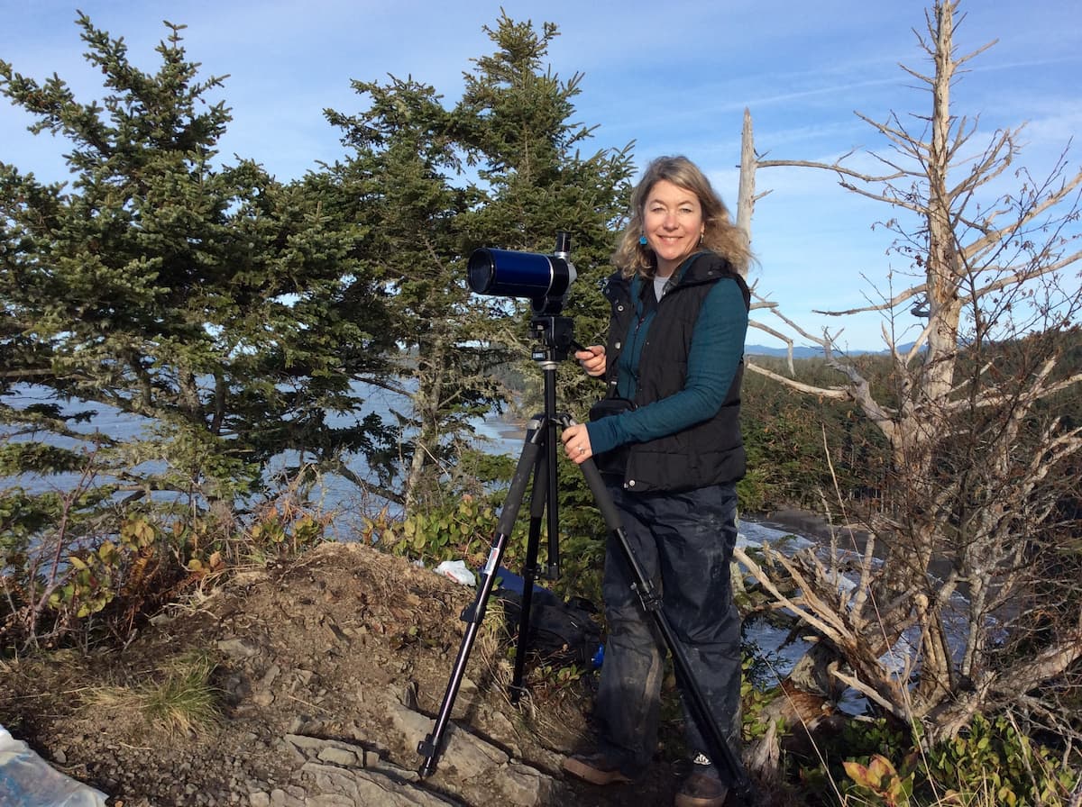 Dr. Shawn Larson standing on a rocky shore and holding a small telescope.