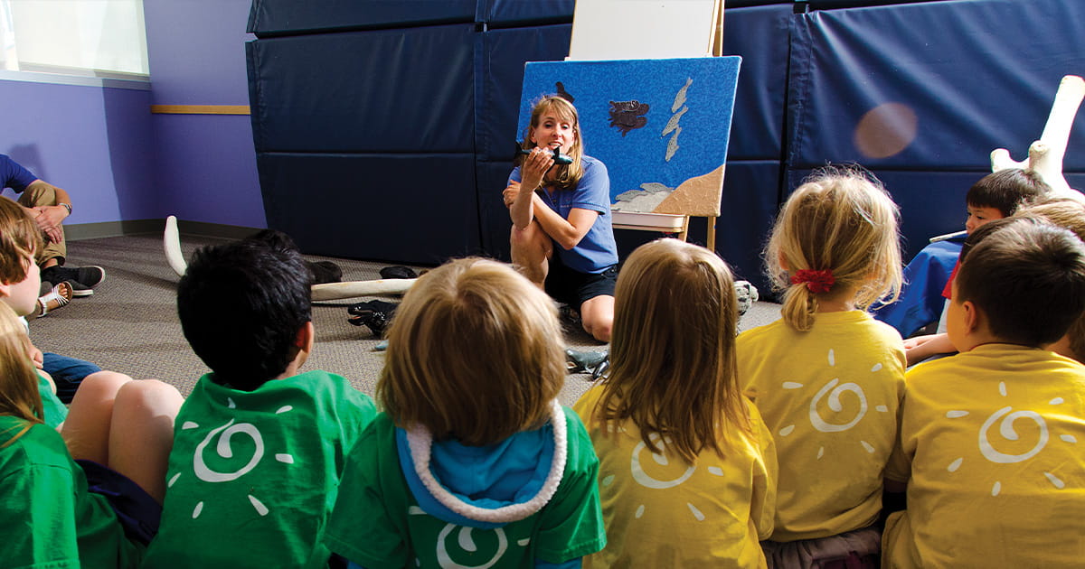 A Seattle Aquarium educator sitting in front of a group of elementary school aged children talking about orca whales while holding a small toy orca.
