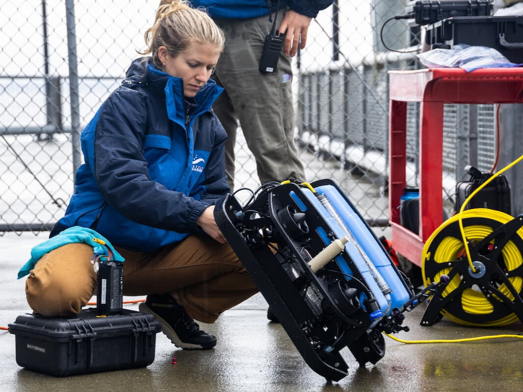 A Seattle Aquarium employee kneeling on a dock. She is holding a blue and black underwater ROV.