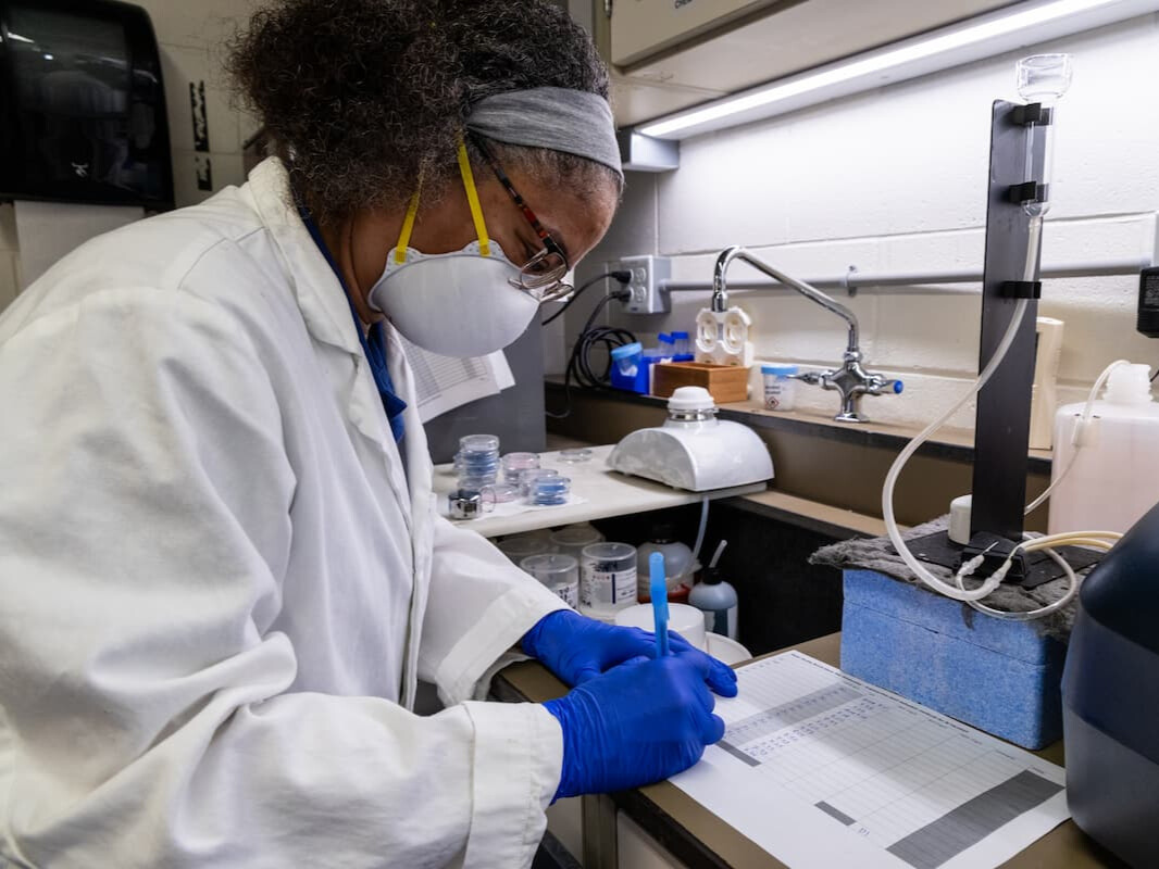 A scientist wearing a white lab coat, blue gloves, and a mask taking notes.