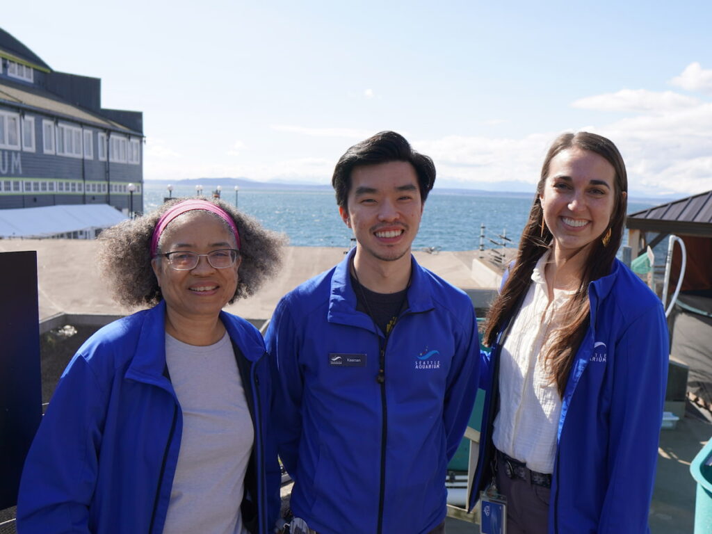 Seattle Aquarium Water Quality team members Angela Smith, Keenan Wong and Hannah Mewhirter standing together and smiling in front of the Salish Sea.
