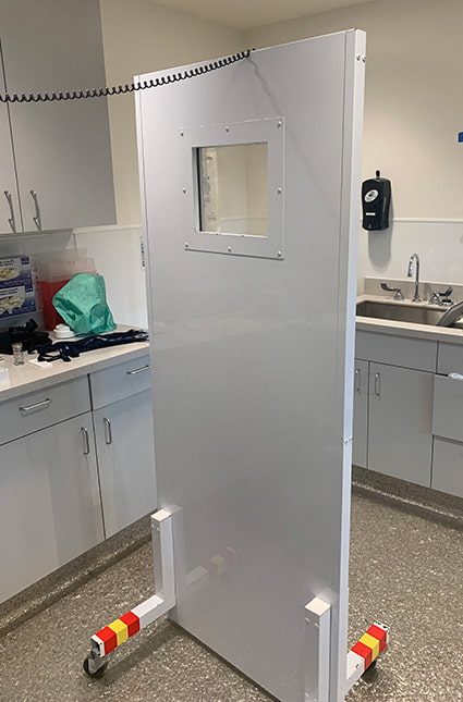 A large lead shield in the shape of a door with a window in the middle with wheels on the bottom positioned inside a bright exam room.