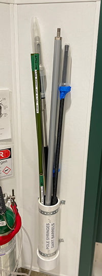 Long poles capable of having syringes attached, all placed inside a white tube which is attached to the wall