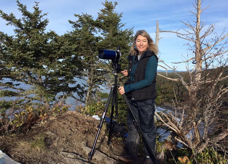 Dr. Shawn Larson standing on a rocky shore and holding a small telescope.