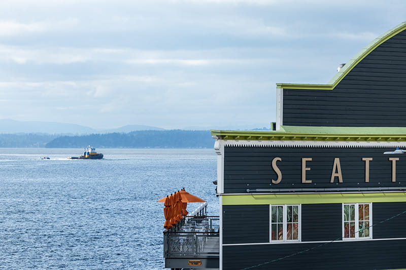 The second level exterior deck of the Seattle Aquarium cafe seen from across the street, against the backdrop of Elliot Bay and a boat moving by in the distance.