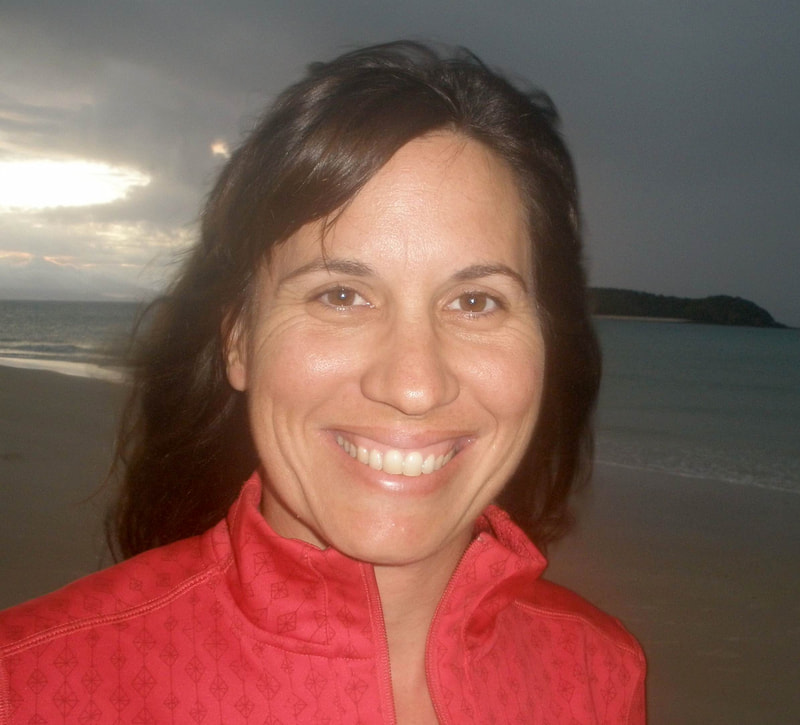 A headshot of Dr. Christine Dudgeon standing in front of the ocean shore. She has long, brown hair and is wearing a red jacket.