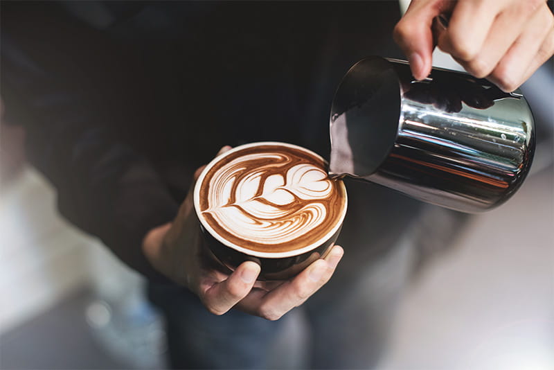 Barista finishing pouring a cup of coffee by adding a foam design on top.