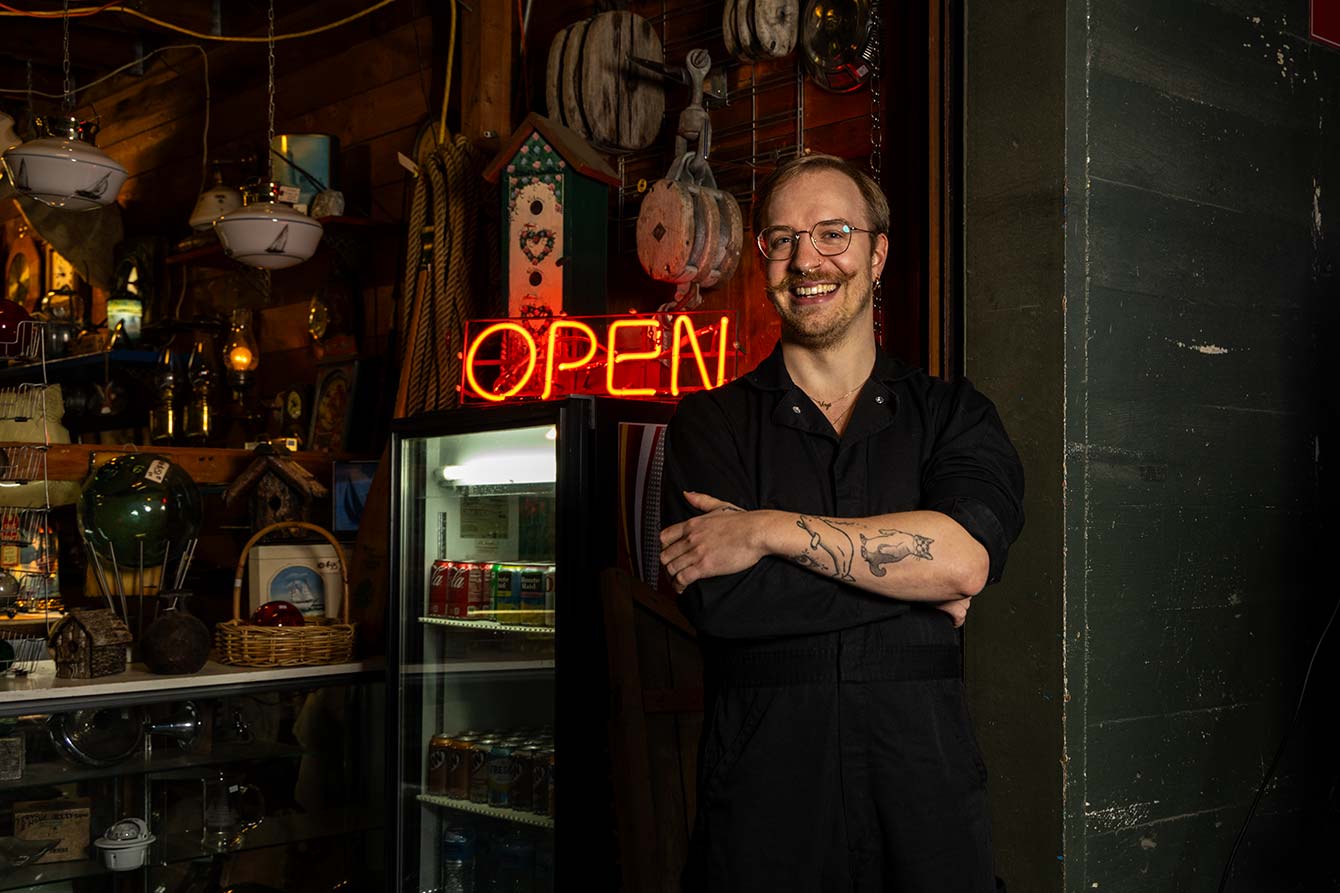 Ben Swenson-Klatt standing in a cluttered antique shop next to a neon red "OPEN" sign. They have short, blonde hair and are wearing glasses and a black jumpsuit. Their arms are crossed, showing tattoos of a cat and a beluga whale.