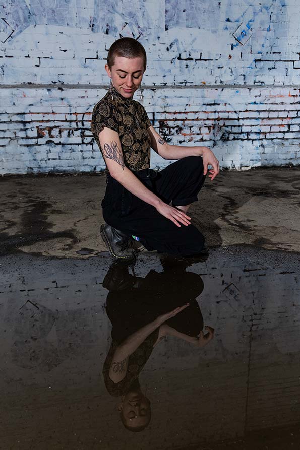 Emma Leiser crouching near a puddle and looking at the reflection of their tattoo in it. Emma has a buzzed head and wears a floral shirt and black pants. Their right arm sports a tattoo of bull kelp.