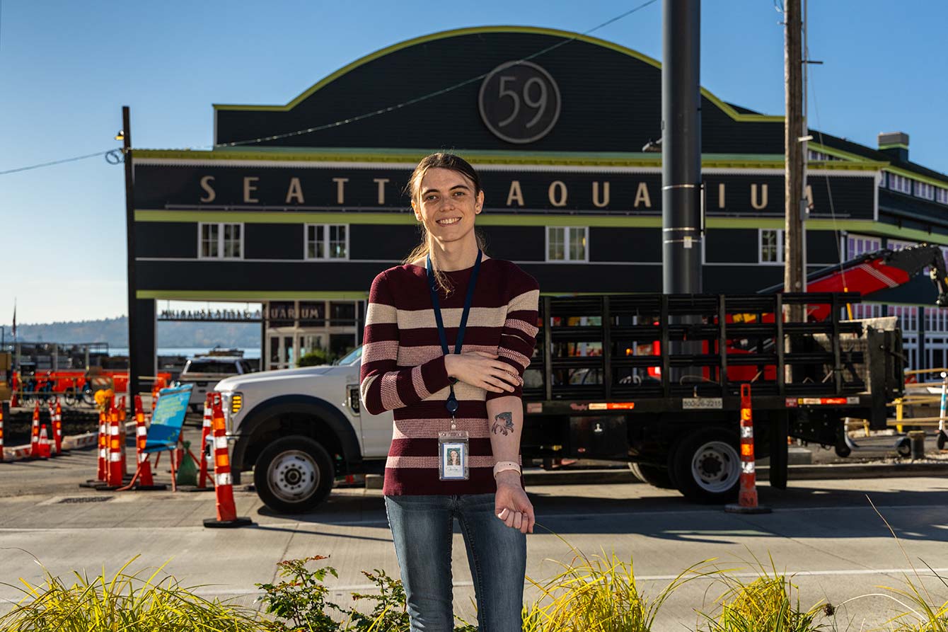 Jessica Williams standing in front of the Seattle Aquarium's entrance. She has long, dark hair in a ponytail and wears a red and white striped shirt and blue jeans. Her left sleeve is partially rolled up to reveal a cuttlefish tattoo on her forearm.