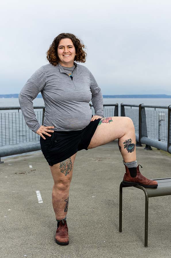 Kaitlin Brawley standing on a pier overlooking the Salish Sea. She has brown, curly hair and wears a grey shirt, black shorts, and brown boots. Her left leg is propped up on a bench, showing off a colorful sunfish tattoo on the inside of her shin.