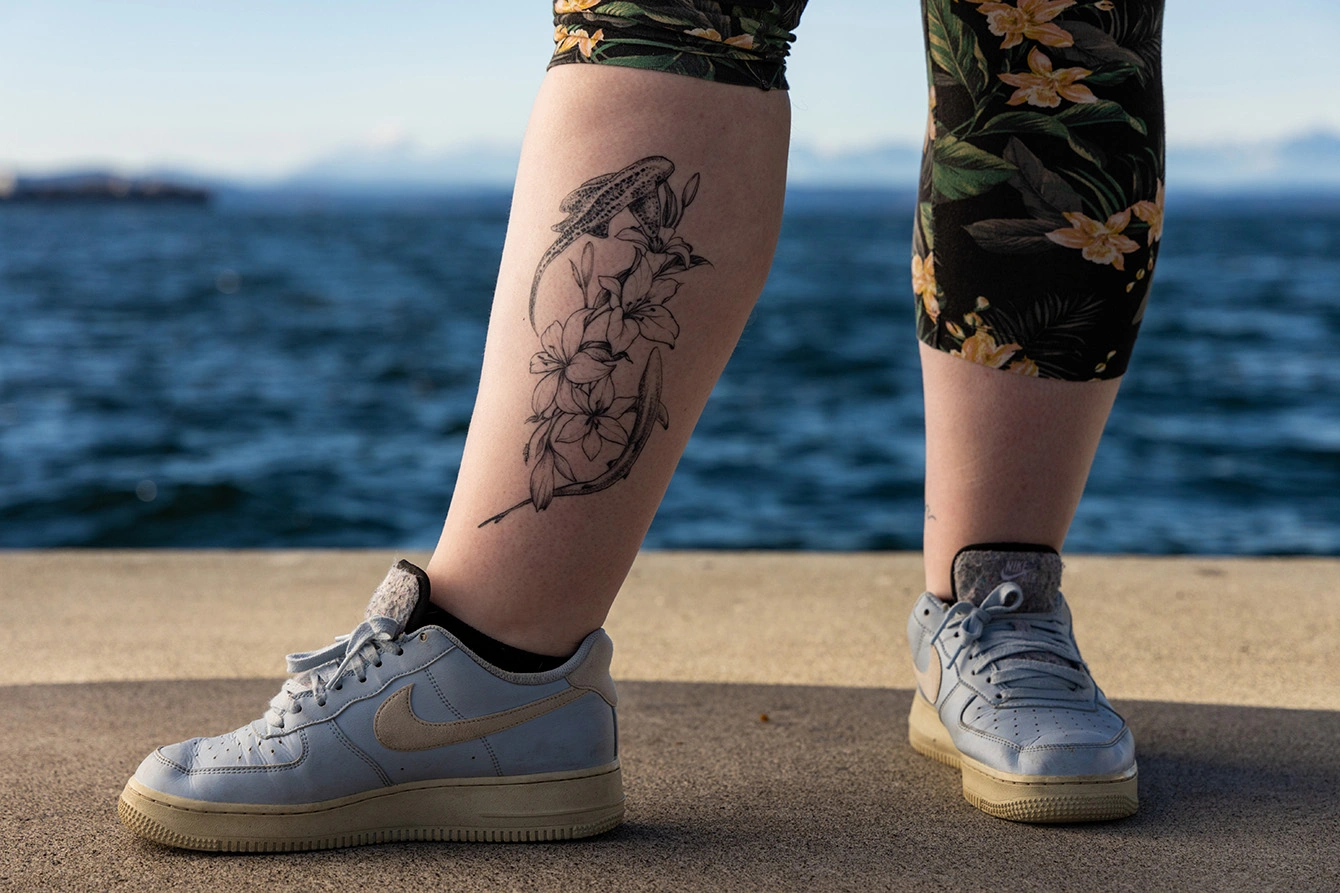 A close-up of Alijah's tattoo on the inside of their right leg. The tattoo depicts an Indo-Pacific leopard shark and a blacktip reef shark swimming around a bundle of white lilies.