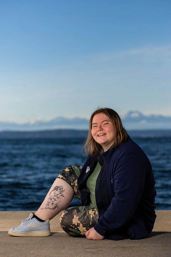Alijah Benbrook sitting on a pier overlooking the Salish Sea. They have shoulder-length blonde hair and are wearing a green shirt, blue jacket, floral pants and light blue tennis shoes. Their right pant leg is rolled up to show a tattoo of two sharks swimming around flowers.