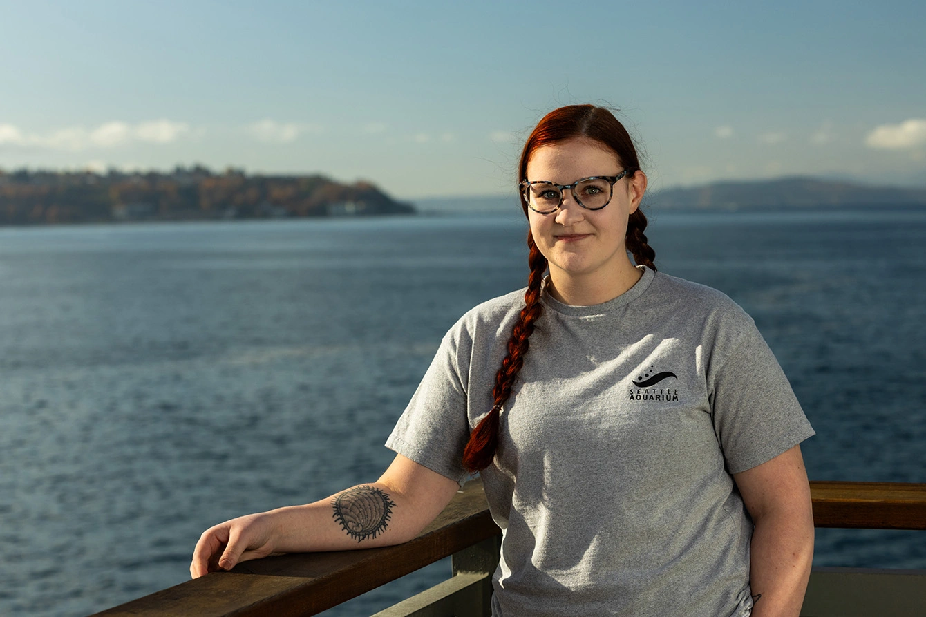 Bailey Johnson standing on a pier overlooking the Salish Sea. She has red hair in two braids, and she wears glasses and a grey t-shirt. A tattoo of a pinto abalone is on her right forearm.