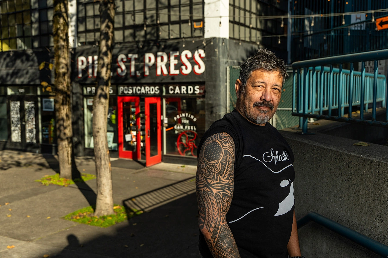Mike Strassburger standing in front of the Pike Street Press storefront. He has short, black hair and wears a black t-shirt. The right sleeve of his shirt is rolled up to reveal a Polynesian sleeve tattoo featuring a sea turtle on his shoulder.