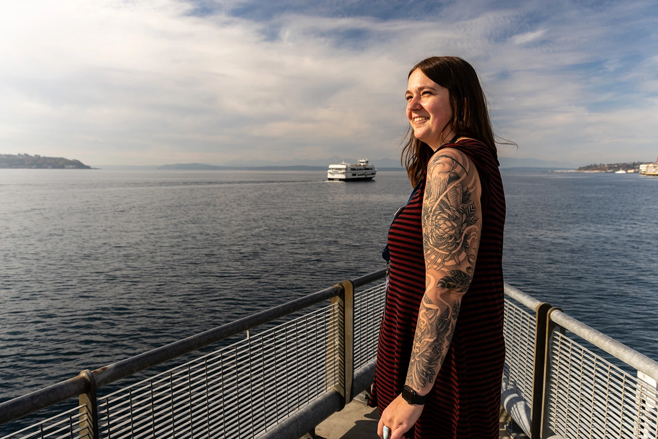 Shelby Kremenich standing on a pier overlooking the Salish Sea. She has long, brown hair and wears a red and black striped dress. Her left sleeve has been rolled up to reveal a sprawling tattoo covering her entire left arm.