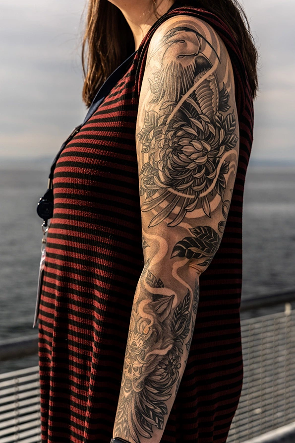 A close-up of Shelby's tattoo on her left arm. Plants and leaves cover her lower arm, and her upper arm sports a blue heron emerging from a bush.