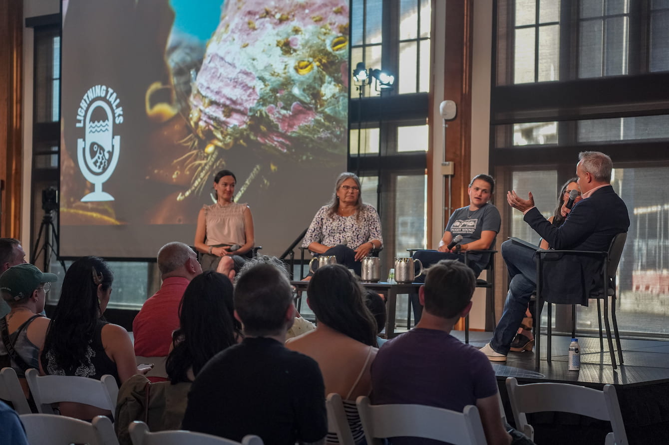 Erin Meyer, Jan Newton, Eileen Bates, Jen Strongin, and Jim Wharton sitting on a low stage in the Seattle Aquarium. Behind them is a projector screen featuring a photo of a pinto abalone and the Lightning Talks logo.