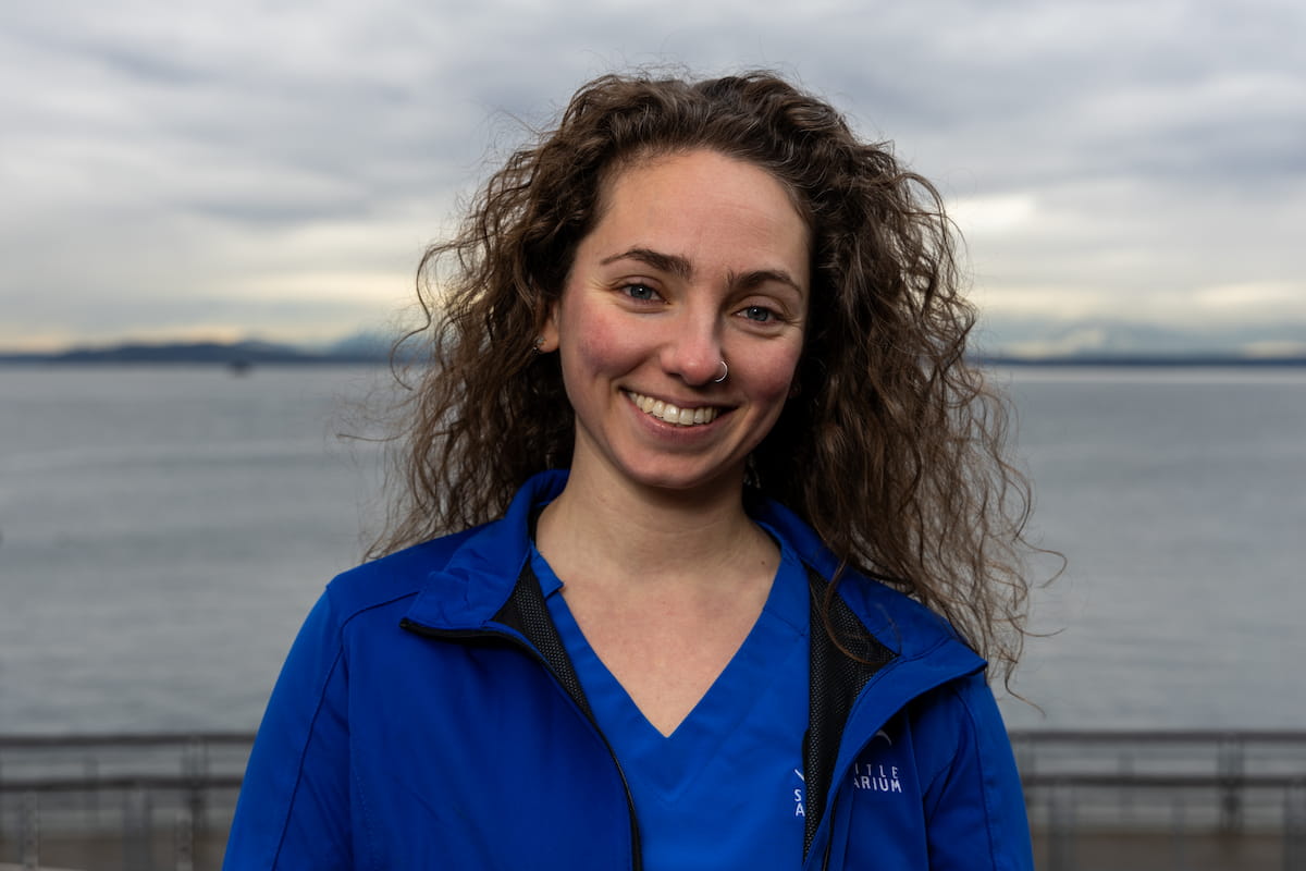 A photo of Dr. Sasha Troiano. She has long, curly brown hair and wears a blue shirt and rain jacket. She is standing on a pier in front of the Puget Sound.