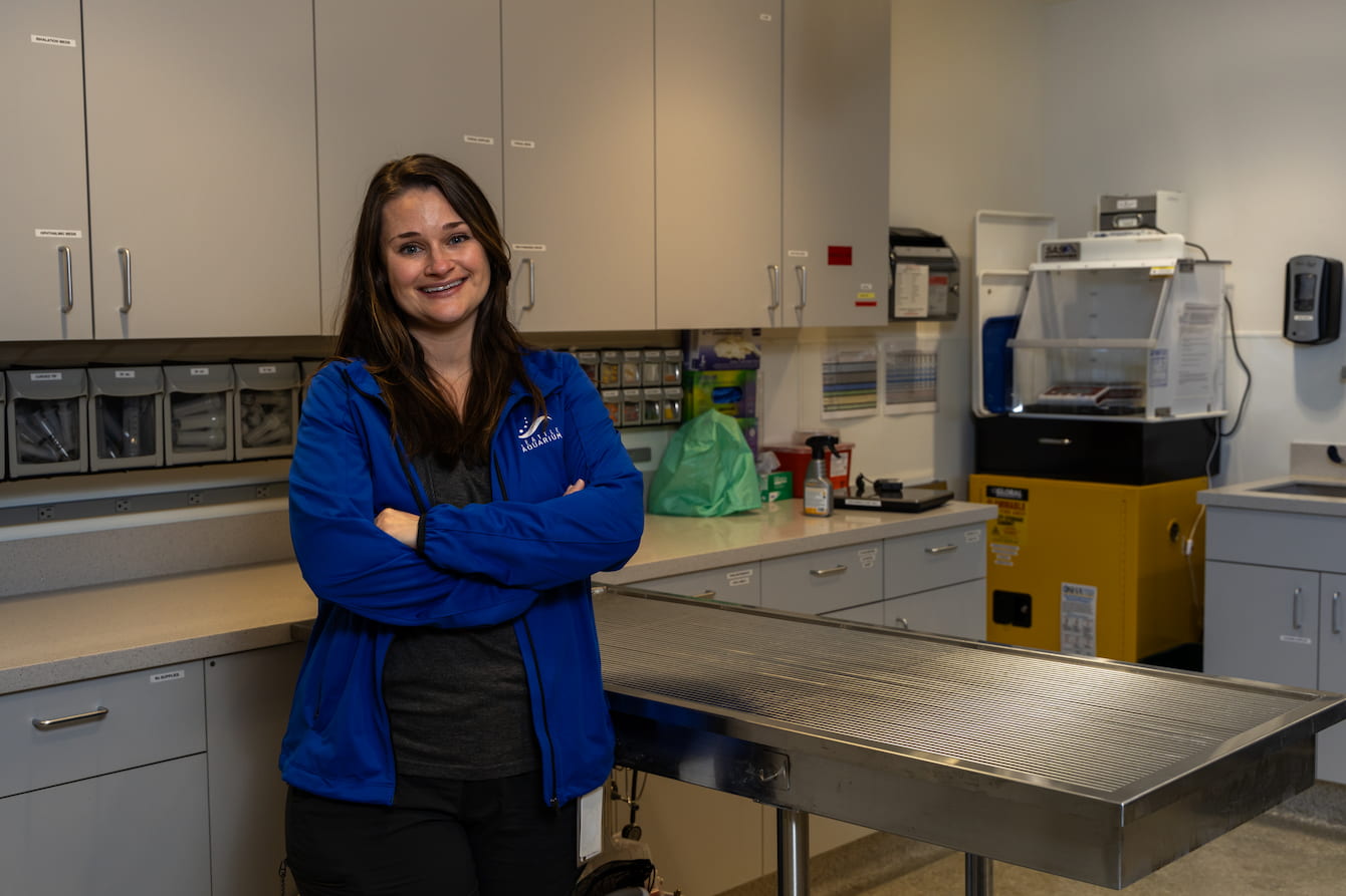 Erika Russ Paz standing in the Seattle Aquarium's Veterinary Care Center. She has straight brown hair and wears a blue Seattle Aquarium jacket.