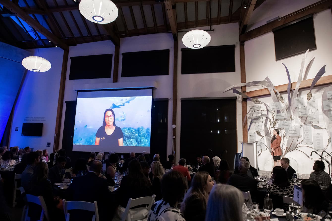 A crowd of seated people viewing Christine Dudgeon as her Zoom call is projected on a large screen.