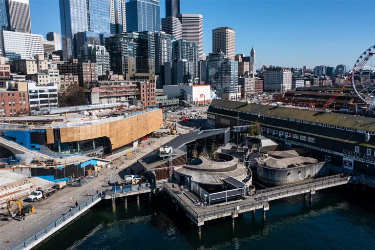 The Seattle Aquarium campus, as seen be drone, looking towards Pier 60, Pier 59 and the under construction Ocean Pavilion with downtown Seattle behind the buildings.