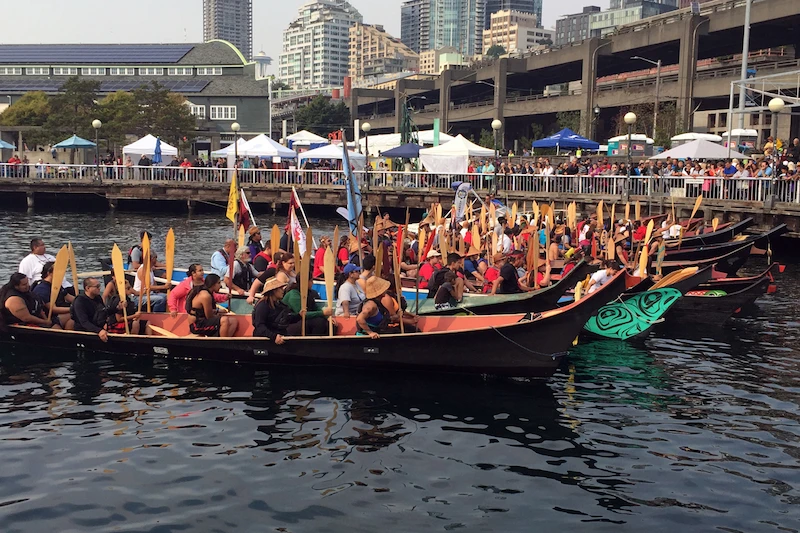 Large canoes filled with individuals holding paddles, part of the Salmon Homecoming event, hosted outside the Seattle Aquarium.