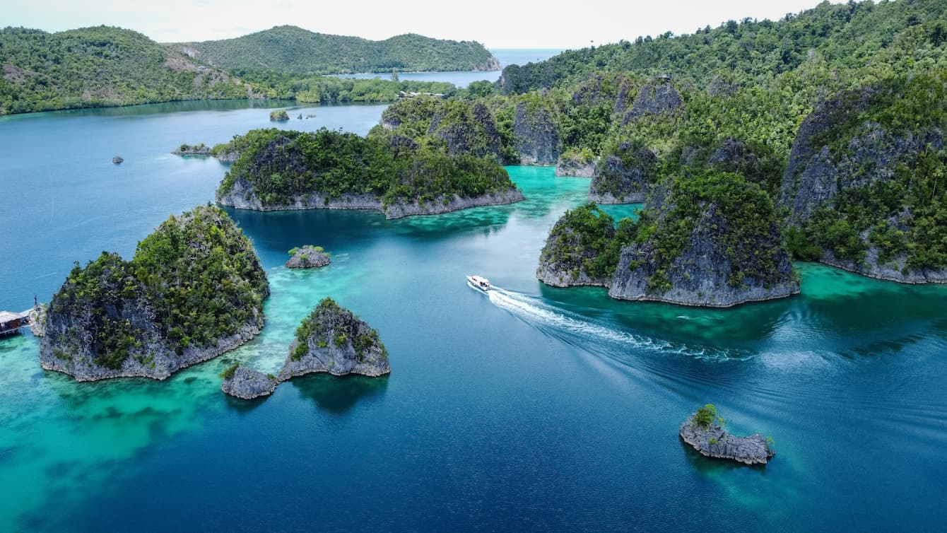 A sweeping drone shot of the waters of the Coral Triangle region. Rocky outcroppings covered in lush vegetation surround the bright blue waters of the sea. A small boat can be seen in the center of the image.