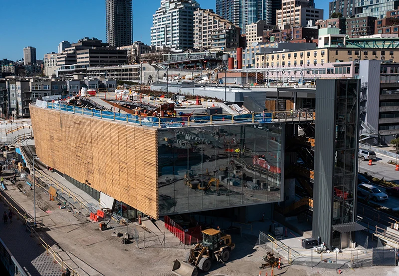 Exterior of the Seattle Aquarium's new Ocean Pavilion building viewed from the front and side, while under construction, showcasing the buildings cedar wood paneling siding and large front glass windows. Construction equipment surrounds the building while work continues on completion.