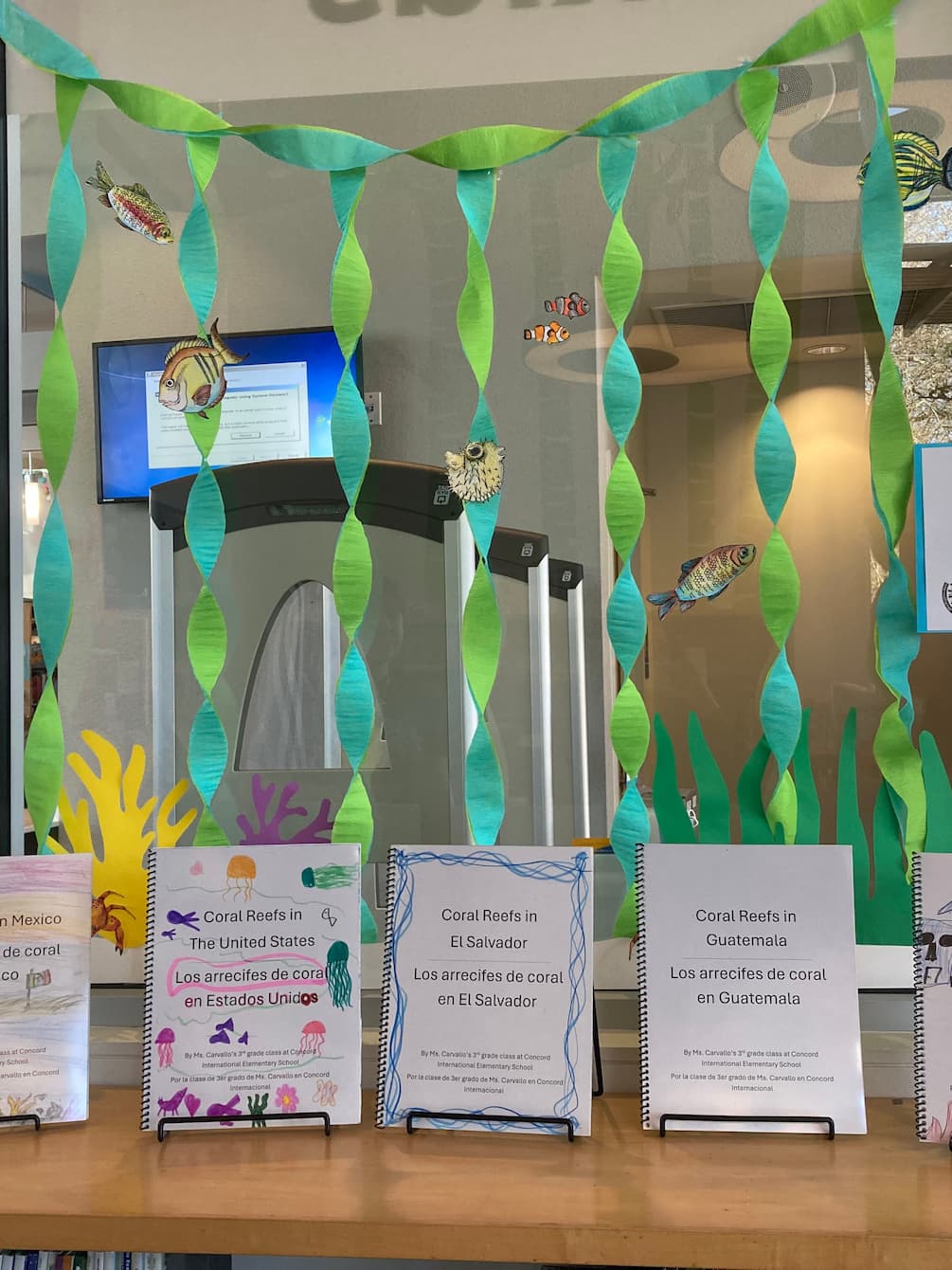The coral reef book exhibit at the South Park library. Several small paper books are lined on a shelf. The space above the shelf is decorated with green and blue streamers, which are meant to resemble kelp, and cut-out illustrations of fish.