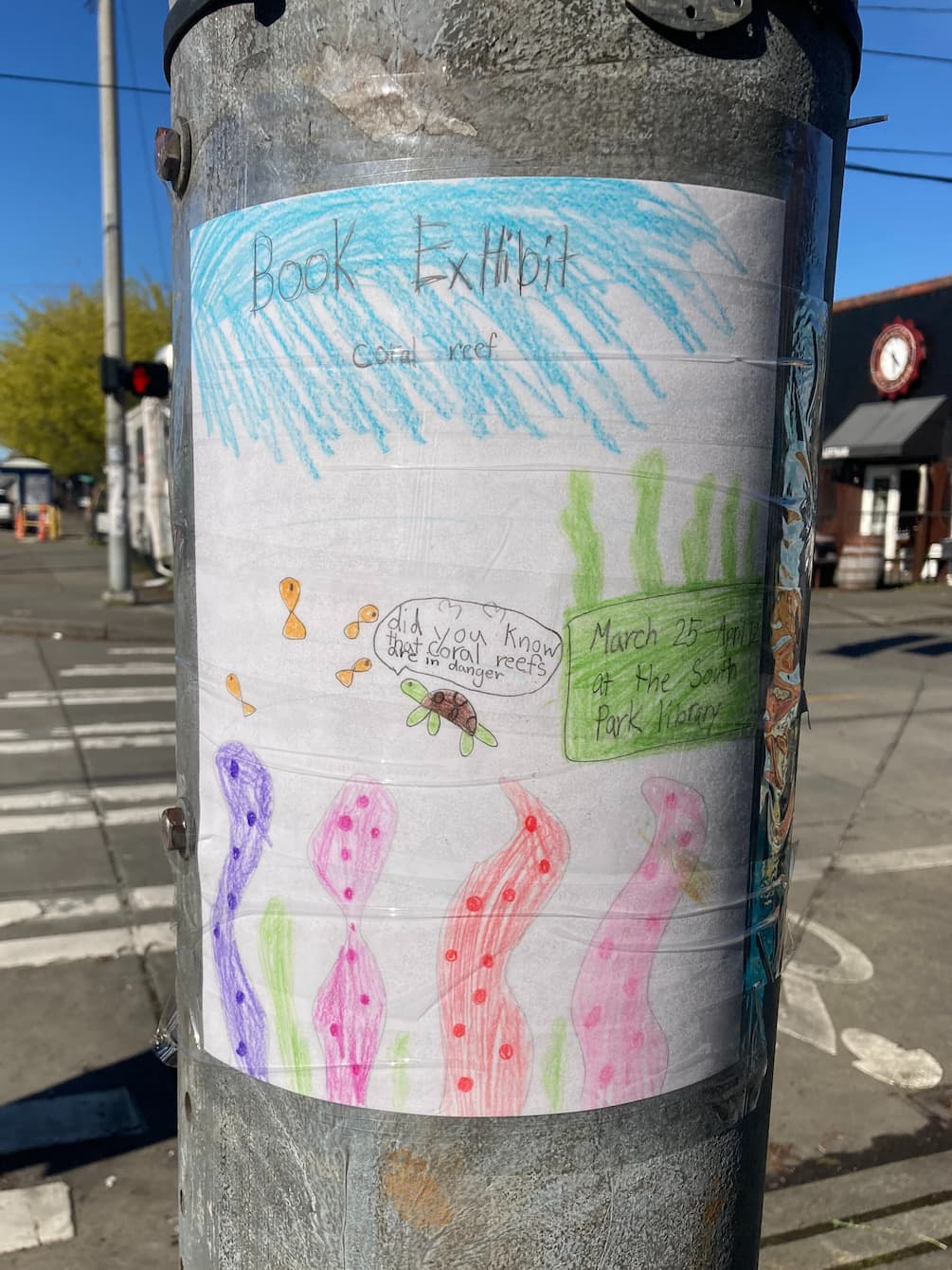 A hand-drawn paper flyer that reads "Book exhibit: coral reef. March 25—April 12 at the South Park library." The flyer has illustrations of coral, kelp, orange fish, and a sea turtle. The sea turtle has a speech bubble over its head that reads "Did you know that coral reefs are in danger."