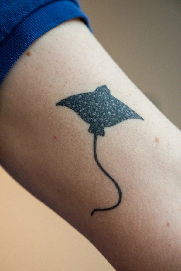 A close-up of Makayla's tattoo: a simple, black illustration of a spotted eagle ray. The tattoo is on the inside of her upper left arm.