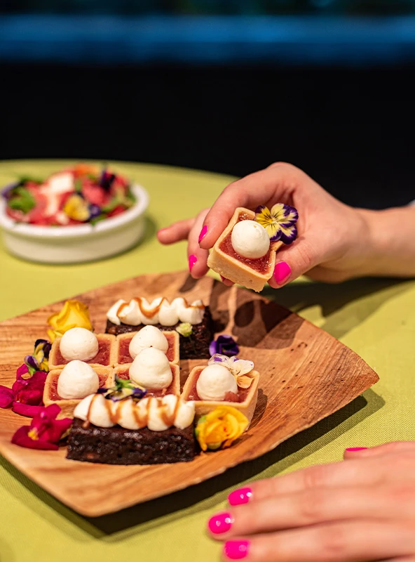 Different desserts placed on a wooden plate which has been decorated with flower petals. An individual has reached to take one of the desserts off the plate with their hand.
