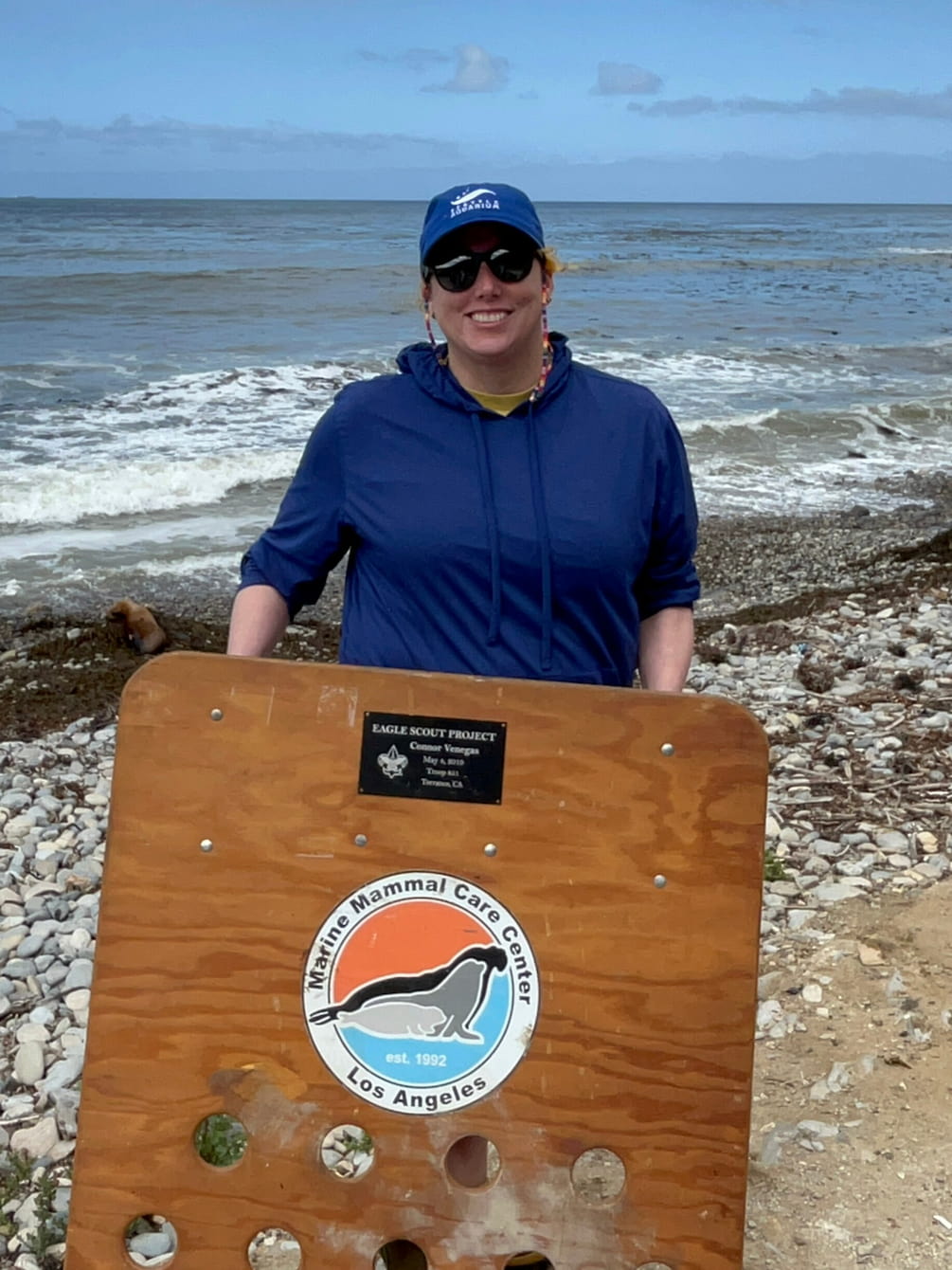 Lindy standing on a beach. She is wearing a blue hat and jacket, and she is holding a wooden herding board.